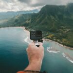 person holding black action camera fronting green mountains